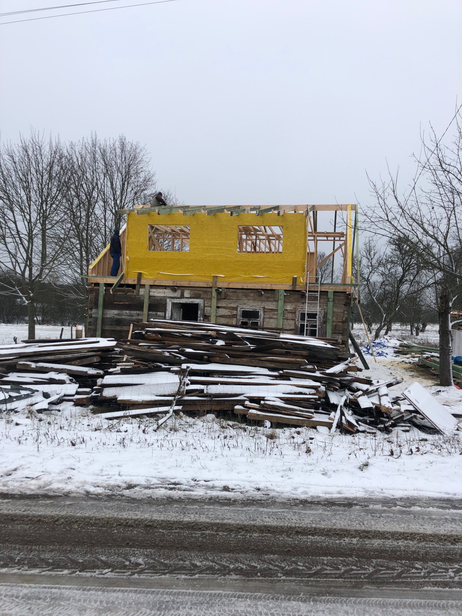 Prace budowlane przy drewnianym domu w zimowej scenerii, widoczne żółte ocieplenie ścian i konstrukcja dachu, na pierwszym planie stos desek pod śniegiem.