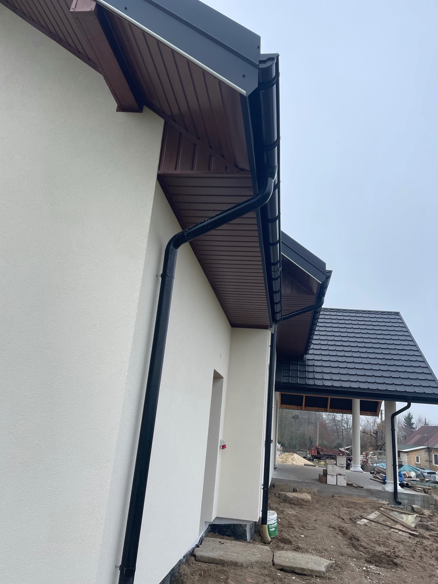 Fragment elewacji domu z zamontowanym systemem rynnowym w kolorze czarnym, widoczna podbitka dachowa w kolorze brązowym i świeżo położony tynk.