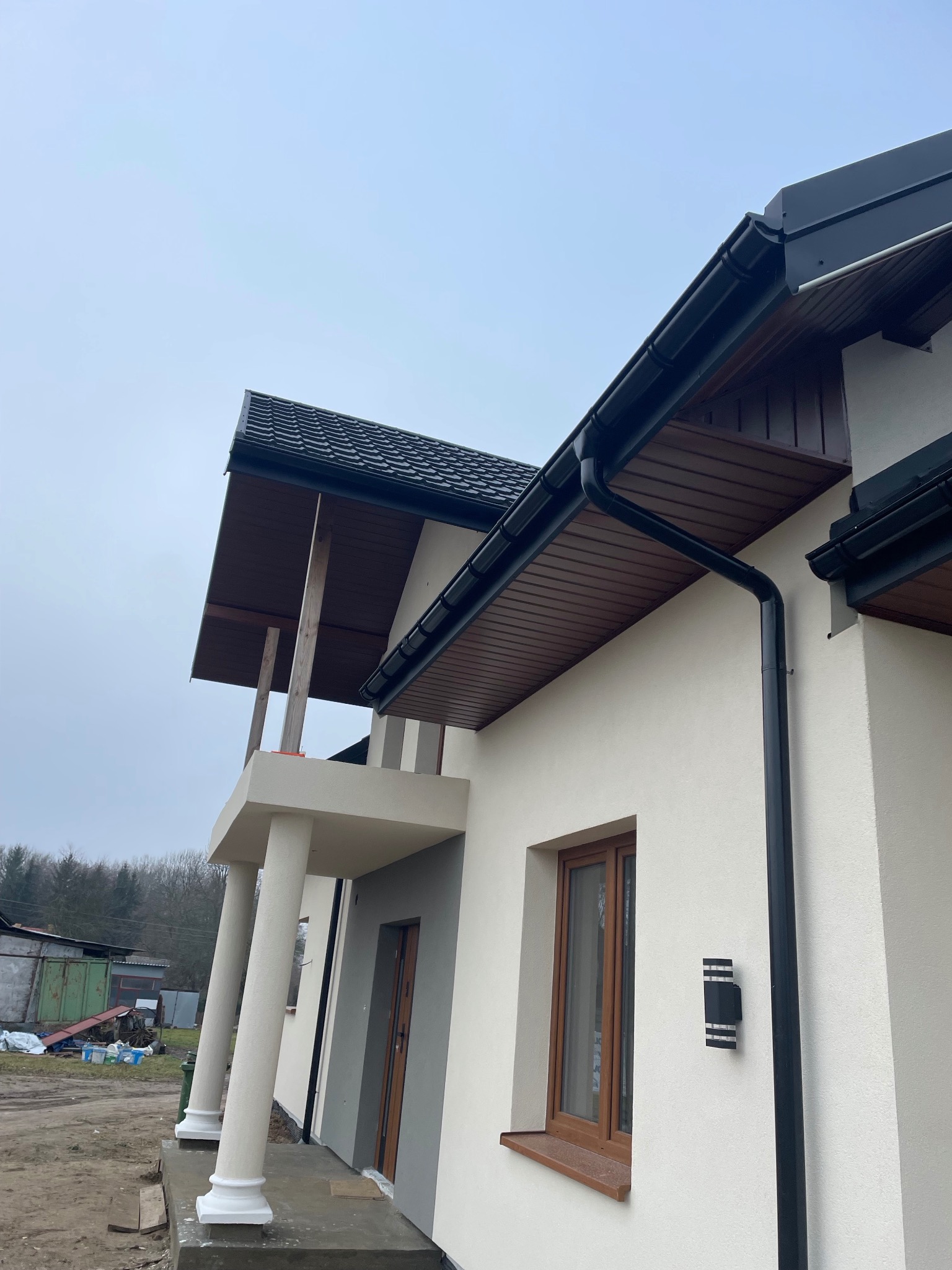 Elewacja domu jednorodzinnego z widocznymi elementami wykończeniowymi: rynny, okna w drewnianej ramie, kolumny podtrzymujące zadaszenie balkonu, zewnętrzna lampa ścienna.