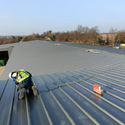 Craft System Sp. z o.o. - Pracownik w kasku i kamizelce odblaskowej montuje blachodachówkę na dachu hali stalowej, widoczny karton z akcesoriami montażowymi.