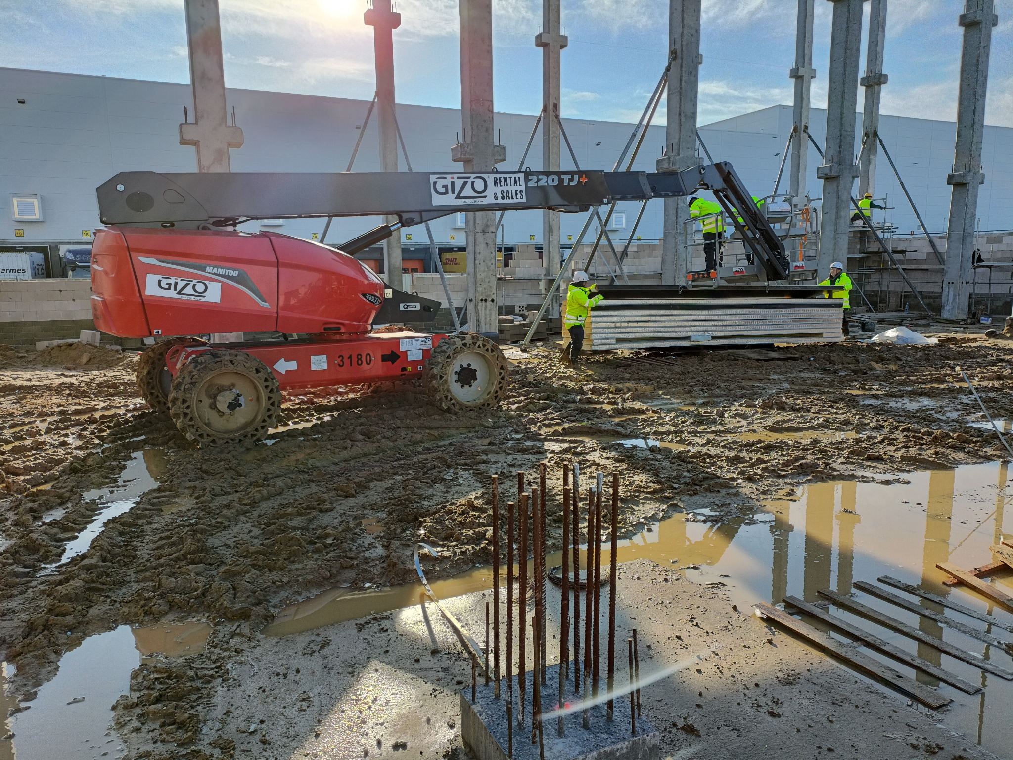Budowa hali: robotnicy w żółtych kamizelkach montują elementy konstrukcyjne z podnośnika koszowego na tle słupów nośnych, widoczna maszyna budowlana Manitou GiZO i błotnisty teren budowy.