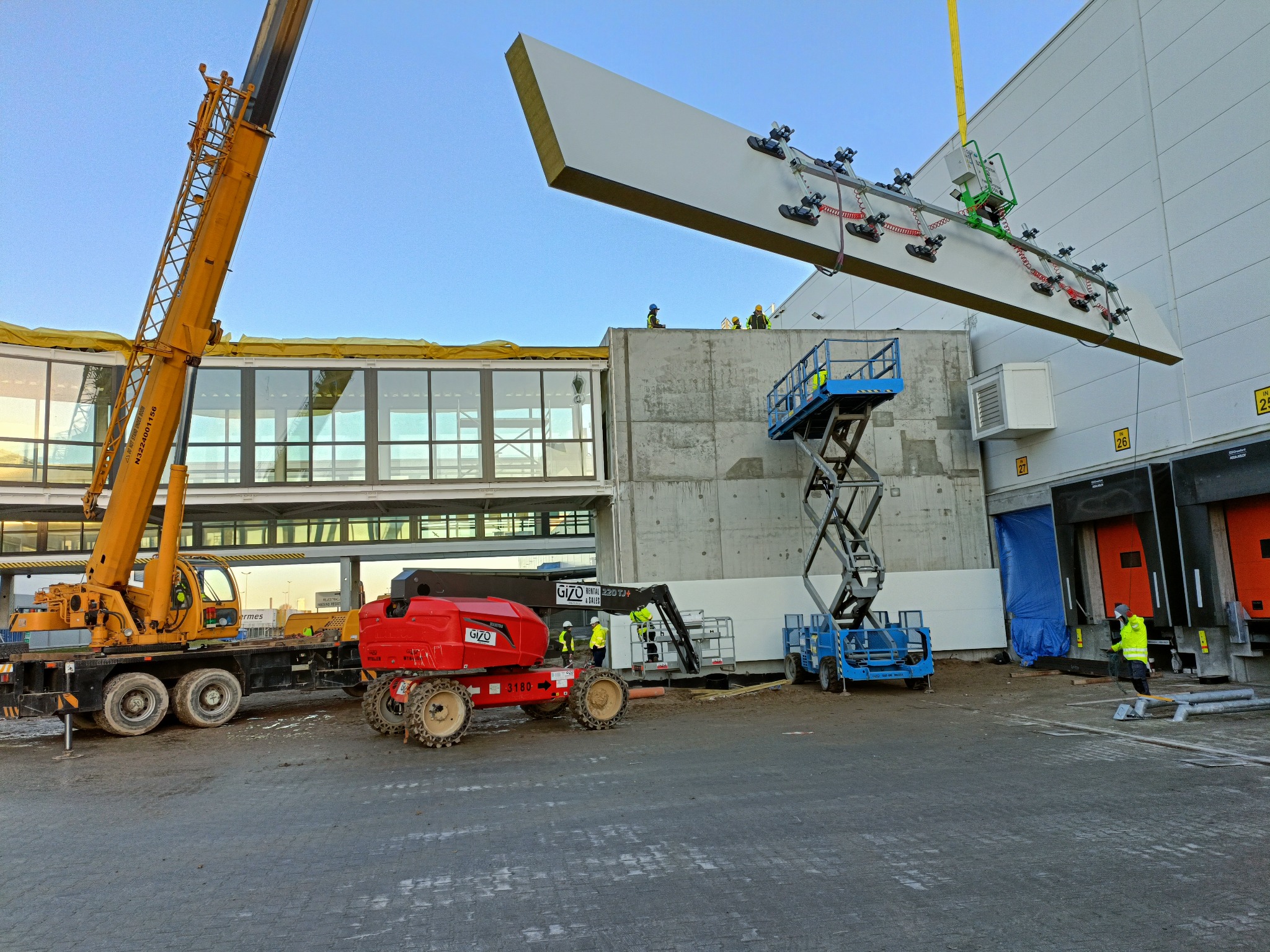 Montaż płyty warstwowej na elewacji hali magazynowej za pomocą przyssawkowej platformy podwieszonej pod dźwigiem, widoczne podnośniki koszowe i pracownicy w odblaskowych kamizelkach.