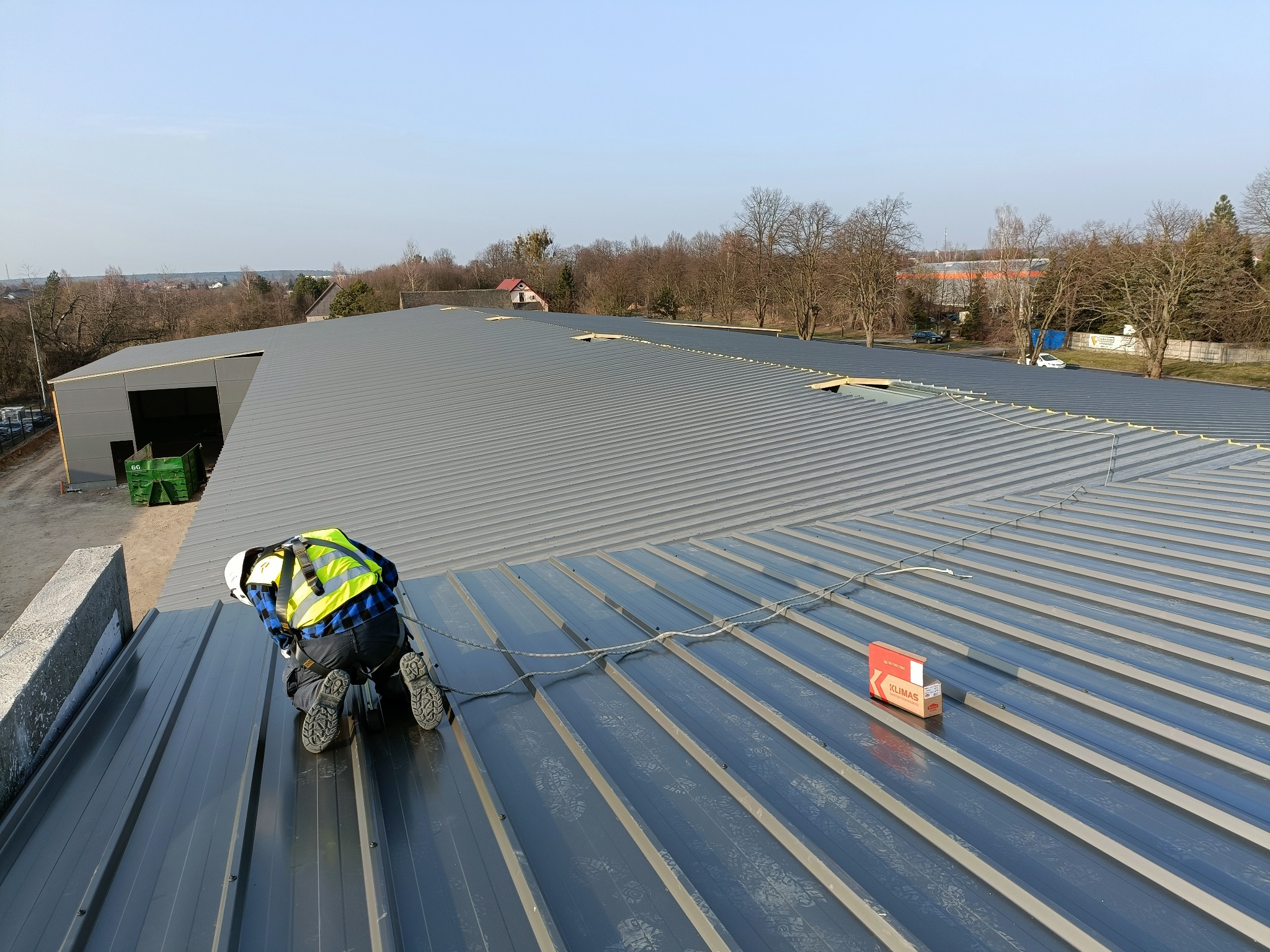 Pracownik w kasku i kamizelce odblaskowej montuje blachodachówkę na dachu hali stalowej, widoczny karton z akcesoriami montażowymi.