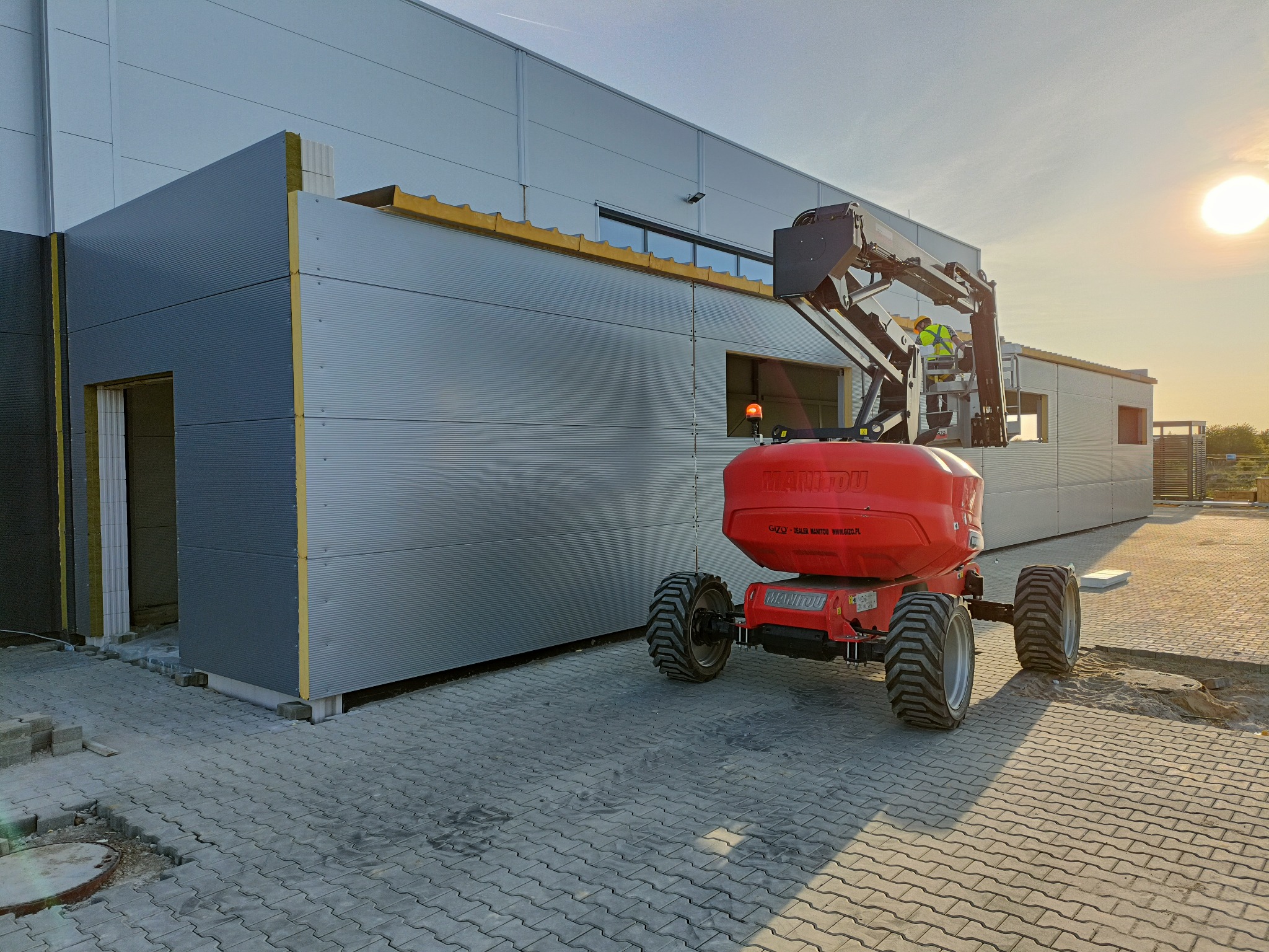 Montaż paneli ściennych na hali przemysłowej z użyciem podnośnika koszowego Manitou, widoczny pracownik w żółtej kamizelce odblaskowej.