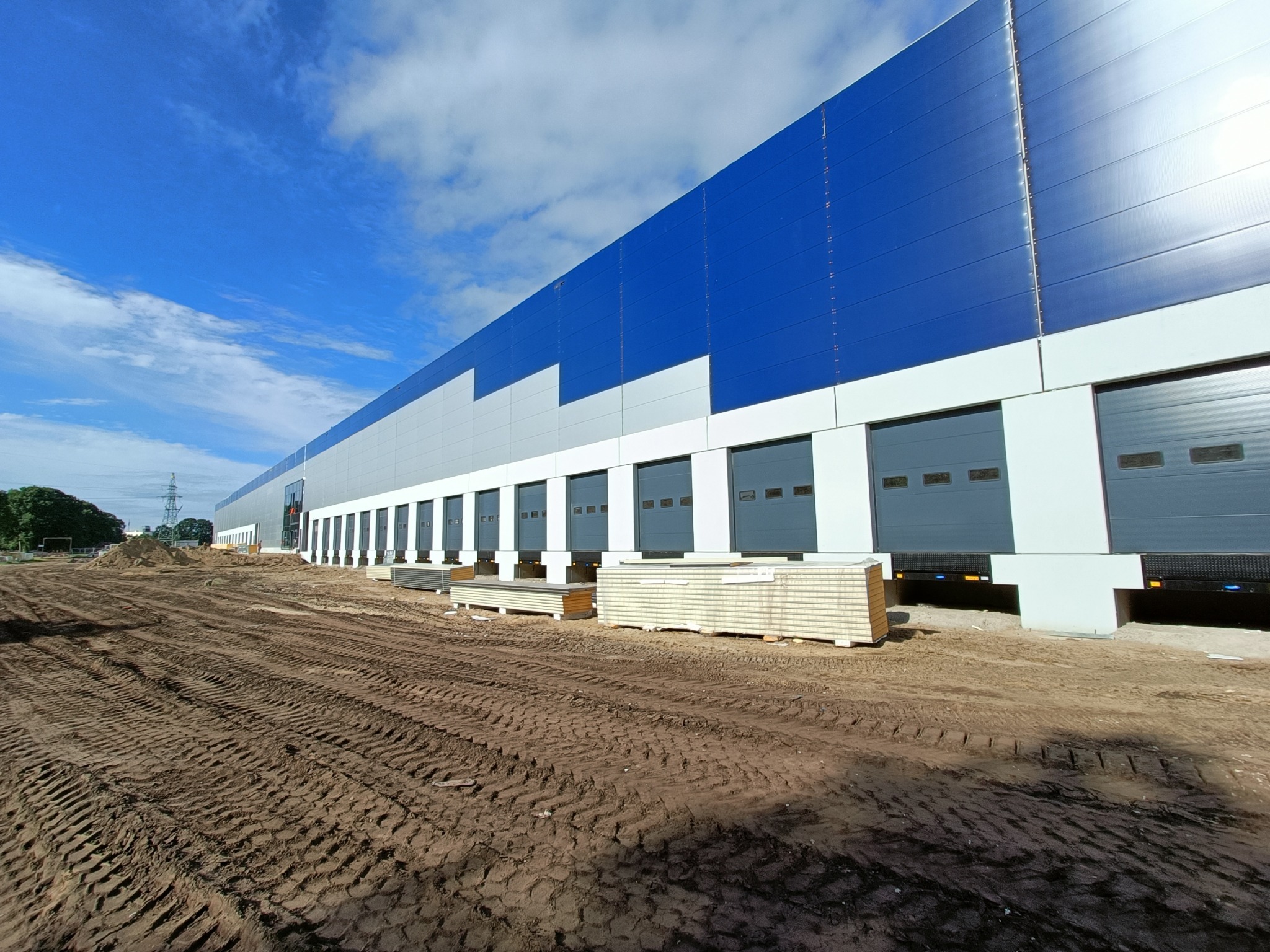 Nowoczesna hala magazynowa z niebiesko-szarą elewacją i sekcyjnymi bramami, widoczna z perspektywy placu budowy z ziemią pooraną śladami opon, pod błękitnym niebem.