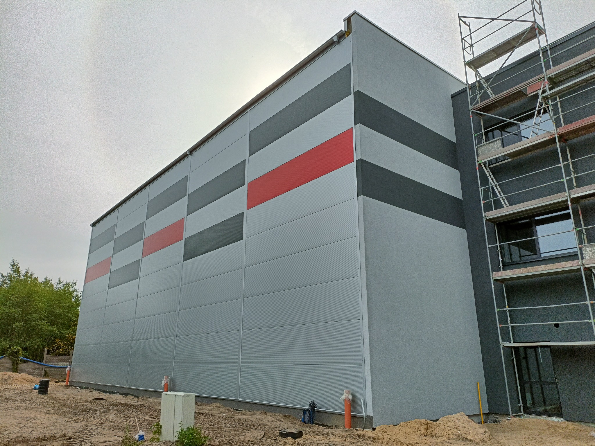 Budynek hali z panelami w odcieniach szarości i czerwieni, widoczne rusztowania i teren budowy.
