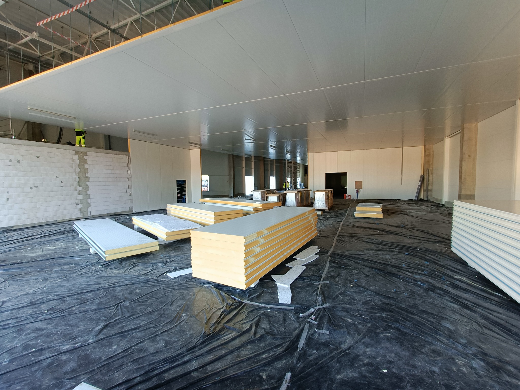 Wnętrze hali w trakcie budowy z ułożonymi na folii panelami warstwowymi, widoczni pracownicy na rusztowaniu i przy ścianie z białego bloczka.