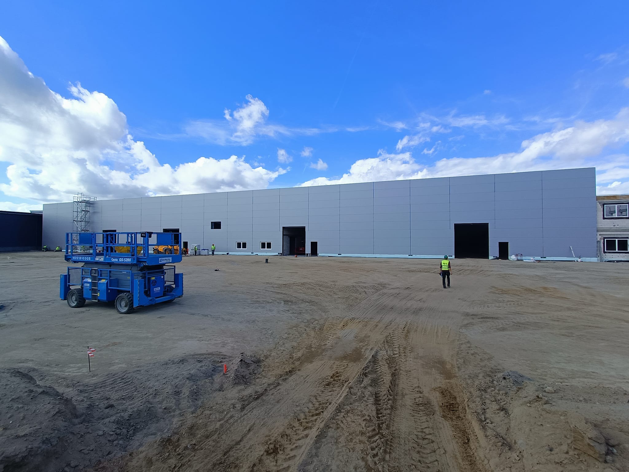 Budowa hali z szarej blachy trapezowej, widoczny podnośnik koszowy i pracownik w kamizelce odblaskowej na placu budowy.