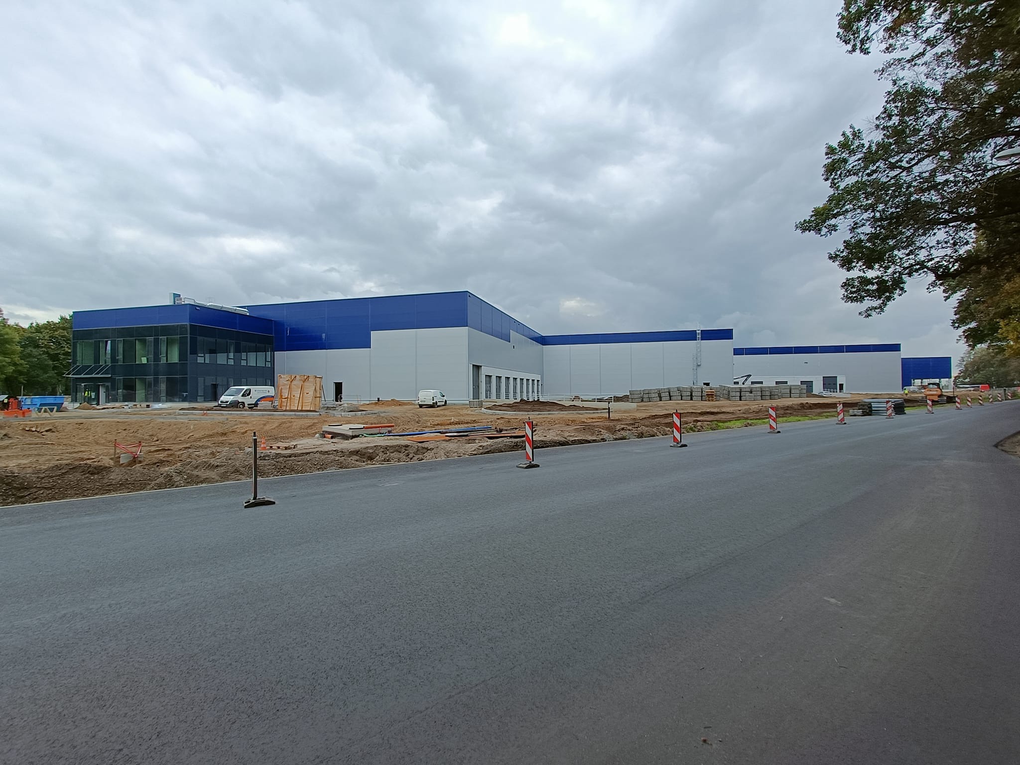 Wznoszona hala produkcyjna z niebieskim dachem i szarą elewacją, widok z drogi, plac budowy z widocznymi materiałami i pojazdami.