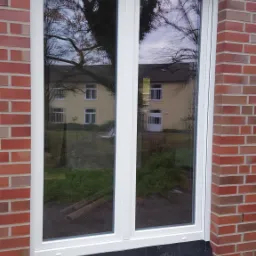 Białe, dwuskrzydłowe okno w nowym murze z czerwonej cegły w Bydgoszczy, z widocznym odbiciem drzewa i budynku w szybie.