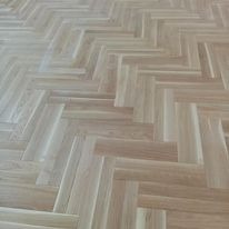 Jasny parkiet ułożony we wzór jodełki francuskiej, widoczne delikatne usłojenie drewna i subtelne refleksy światła.