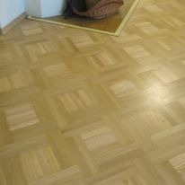 Jasna, lakierowana podłoga z parkietu ułożonego we wzór kwadratów z przesunięciem, widoczny narożnik ściany i fragment koszyka w tle.