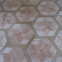 Parkiet ułożony w sześciokątną mozaikę z wzorem rozety w centrum każdego elementu, wykonany z jasnego drewna z wyraźnym usłojeniem, widoczne niewielkie otwory po gwoździach.