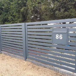 Nowoczesne, poziome, szare ogrodzenie panelowe z zamontowaną skrzynką pocztową z numerem 86, na tle zielonych drzew iglastych.