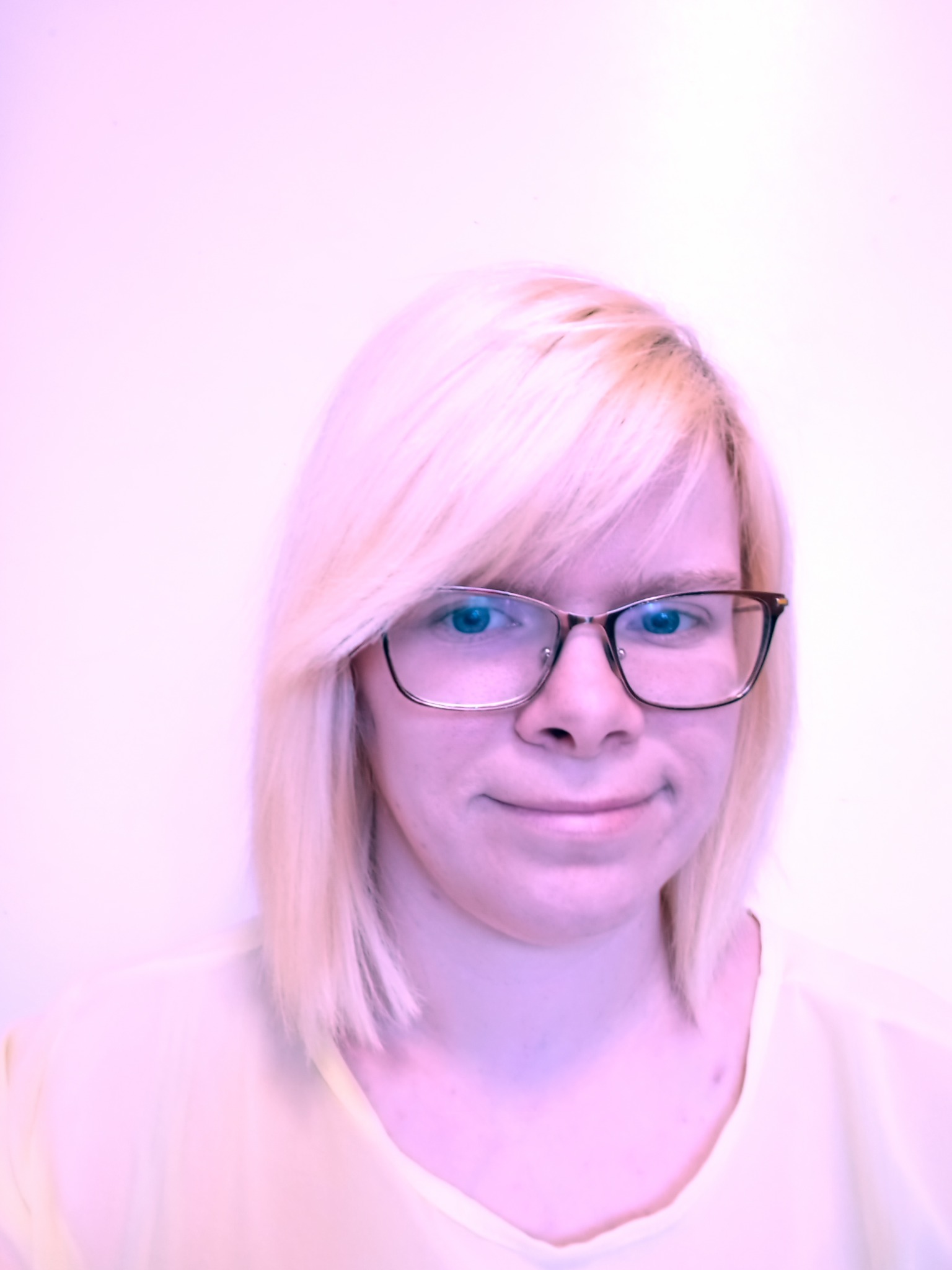 Portret kobiety z blond włosami i okularami o metalowych oprawkach, uśmiechającej się delikatnie na jasnym, rozmytym tle.