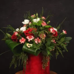 Kompozycja kwiatowa w czerwonym, cylindrycznym wazonie na drewnianym stołku, zawierająca czerwone i białe róże, goździki z czerwonym obrzeżem, zielone chryzantemy oraz gałązki iglaste, ozdobiona...