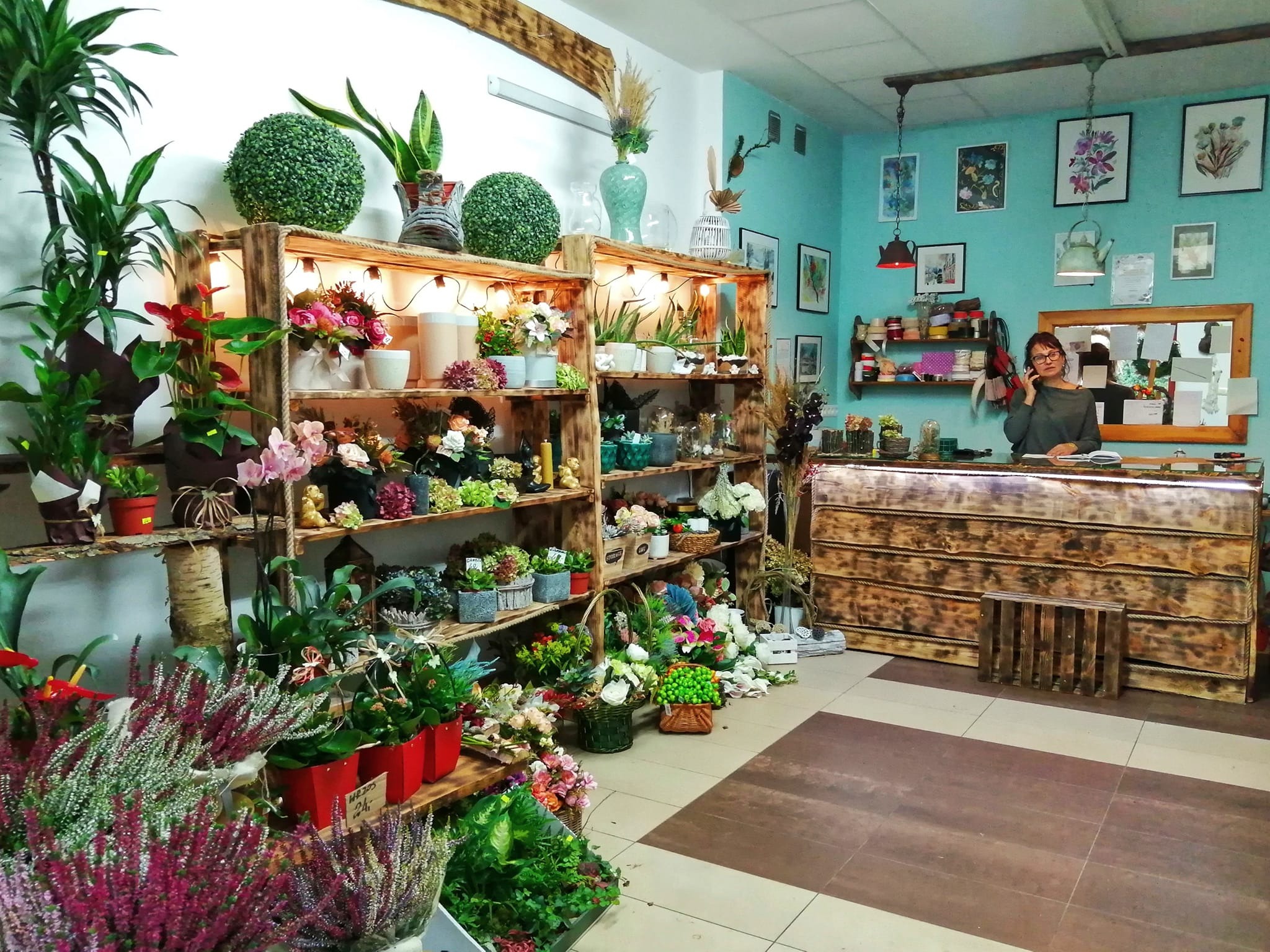 Wnętrze kwiaciarni w Suwałkach z drewnianymi regałami pełnymi różnorodnych roślin doniczkowych, kwiatów ciętych i dekoracji, za ladą stoi kobieta rozmawiająca przez telefon.