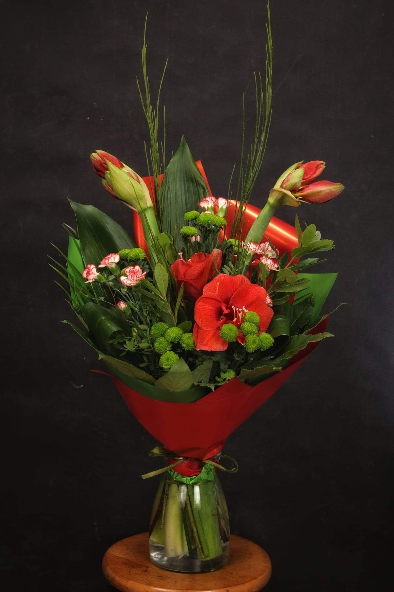 Bukiet z czerwonymi amarylisami, goździkami i zielonymi santini, owinięty w czerwony papier i umieszczony w szklanym wazonie na tle ciemnej ściany.