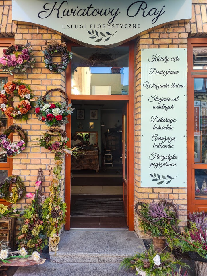 Wejście do kwiaciarni 'Kwiatowy Raj' z ceglaną fasadą, ozdobionej różnorodnymi wieńcami kwiatowymi i kompozycjami roślinnymi, z tablicą informacyjną o usługach florystycznych.