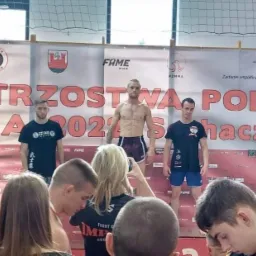 Zawodnicy na podium podczas zawodów sportów walki, widoczny plakat 'Mistrzostwa Polski 2023', publiczność obserwuje.