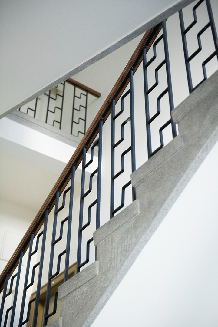 Schody betonowe z drewnianą poręczą i metalową balustradą o geometrycznym wzorze, widziane z dołu pod kątem, z fragmentem drugiego biegu schodów w tle.