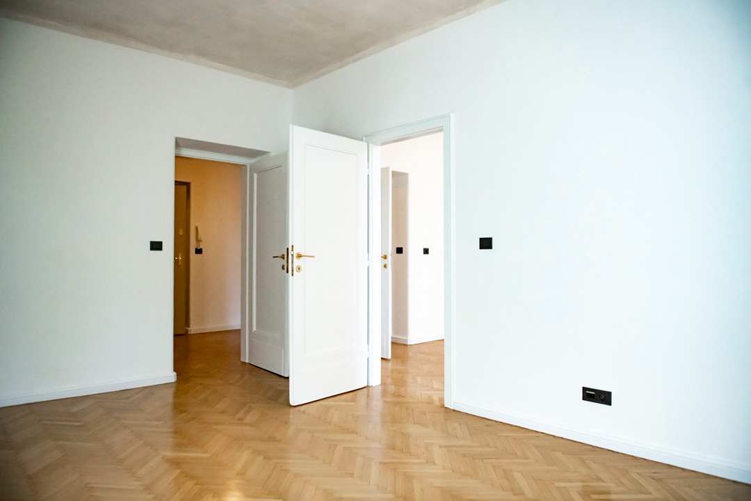 Puste, jasne wnętrze z parkietem w jodełkę, białymi ścianami i otwartymi białymi drzwiami z mosiężnymi klamkami, widoczne czarne gniazdka elektryczne.