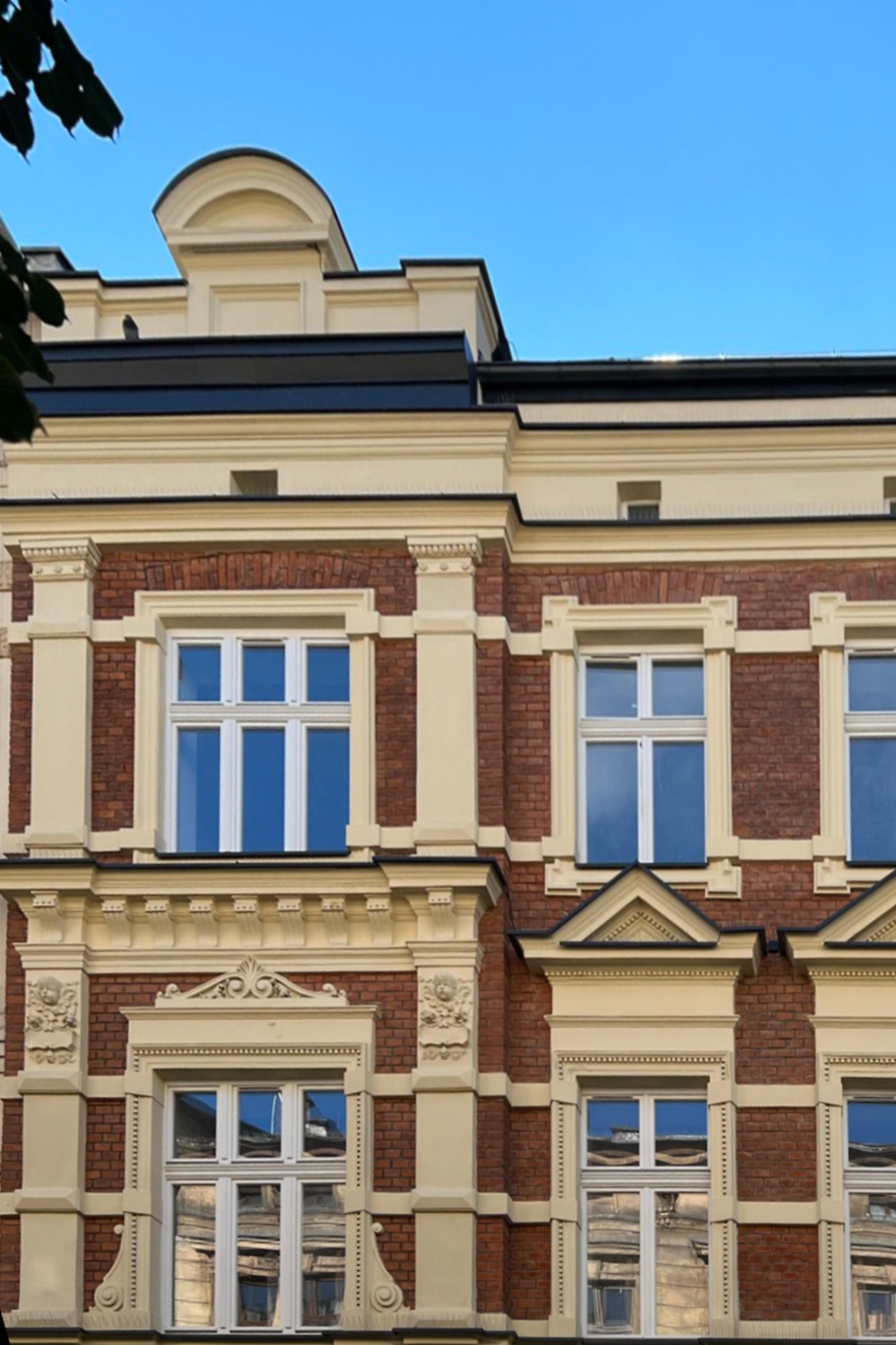 Fasada kamienicy z cegły i kremowymi zdobieniami, okna z refleksami nieba, widok z dołu na błękitne niebo.