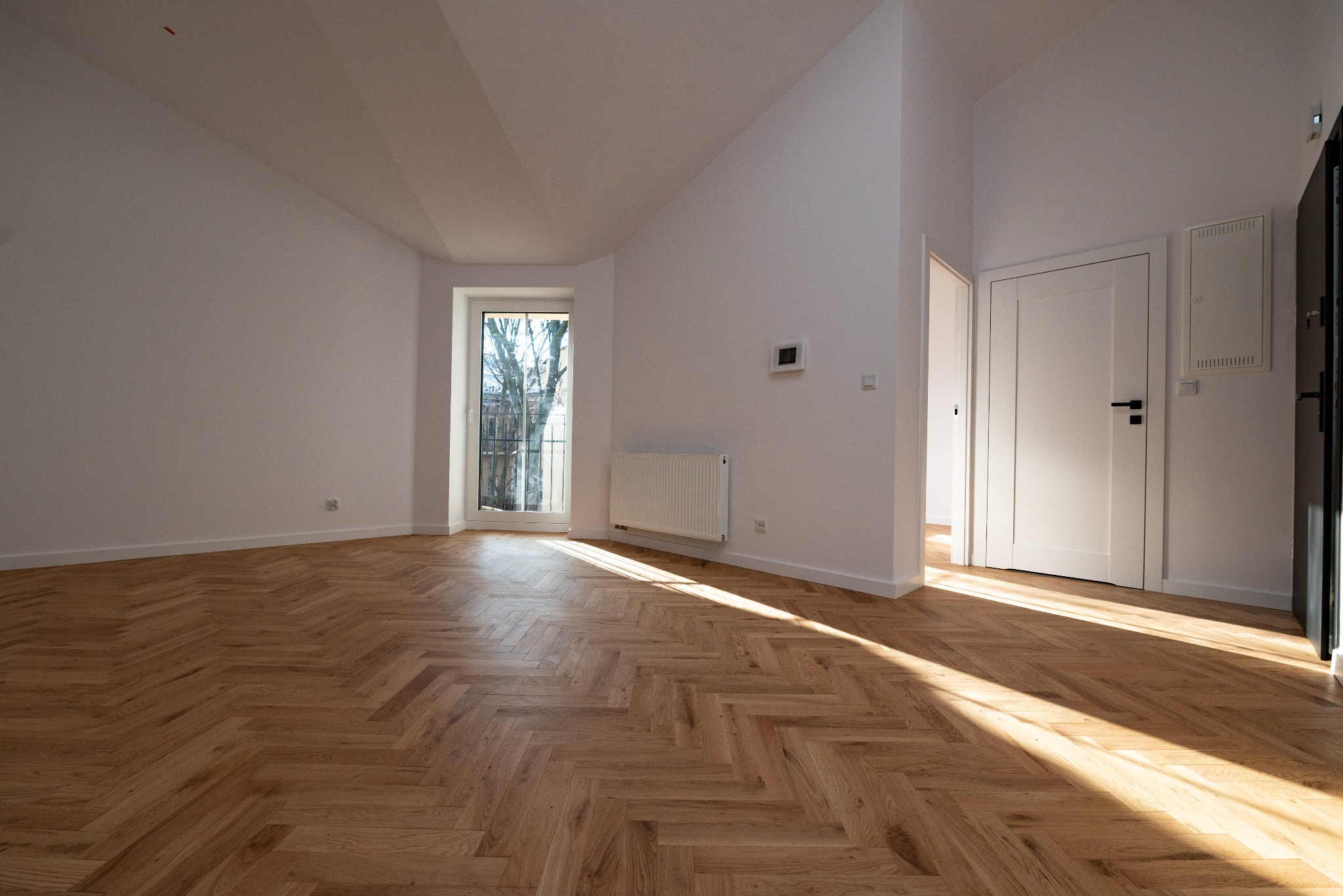 Pusty, jasny pokój z parkietem ułożonym w jodełkę, białymi ścianami i drzwiami, widok z okna na drzewa, grzejnik pod oknem, wpadające promienie słoneczne.