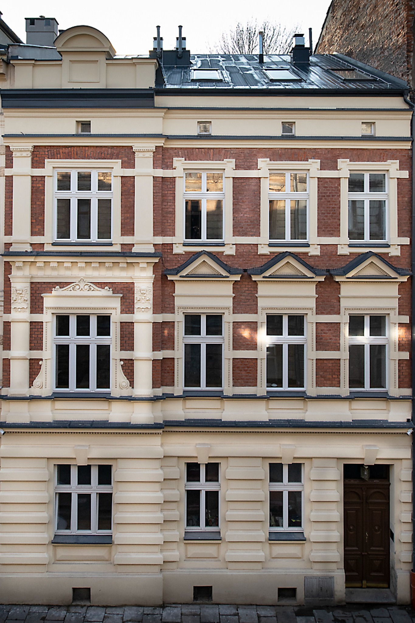 Fasada kamienicy z czerwonej cegły z jasnymi elementami dekoracyjnymi, widoczne okna z białymi ramami i drewniane drzwi wejściowe.