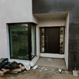 Nowoczesny dom w trakcie wykończenia z zamontowanymi oknami z czarną ramą i ciemnymi drzwiami wejściowymi, widoczne materiały budowlane i narzędzia.