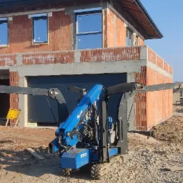 Niedokończony dom jednorodzinny z zamontowanymi oknami i bramą garażową, widziany z perspektywy małego dźwigu budowlanego na gąsienicach.