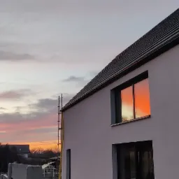 Nowoczesny dom jednorodzinny z czarną dachówką i oknami, w których odbija się pomarańczowy zachód słońca, na tle zabudowy sąsiedzkiej i pochmurnego nieba.