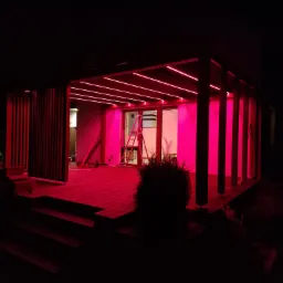 Nowoczesna pergola z drewnianym tarasem oświetlona intensywnym różowym światłem LED, widoczna drabina oparta o ścianę, nocne ujęcie.