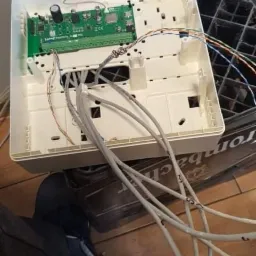 Otwarta biała skrzynka z zieloną płytką drukowaną i podłączonymi szarymi kablami na drewnianej podłodze, obok czarnej skrzynki z tworzywa sztucznego.