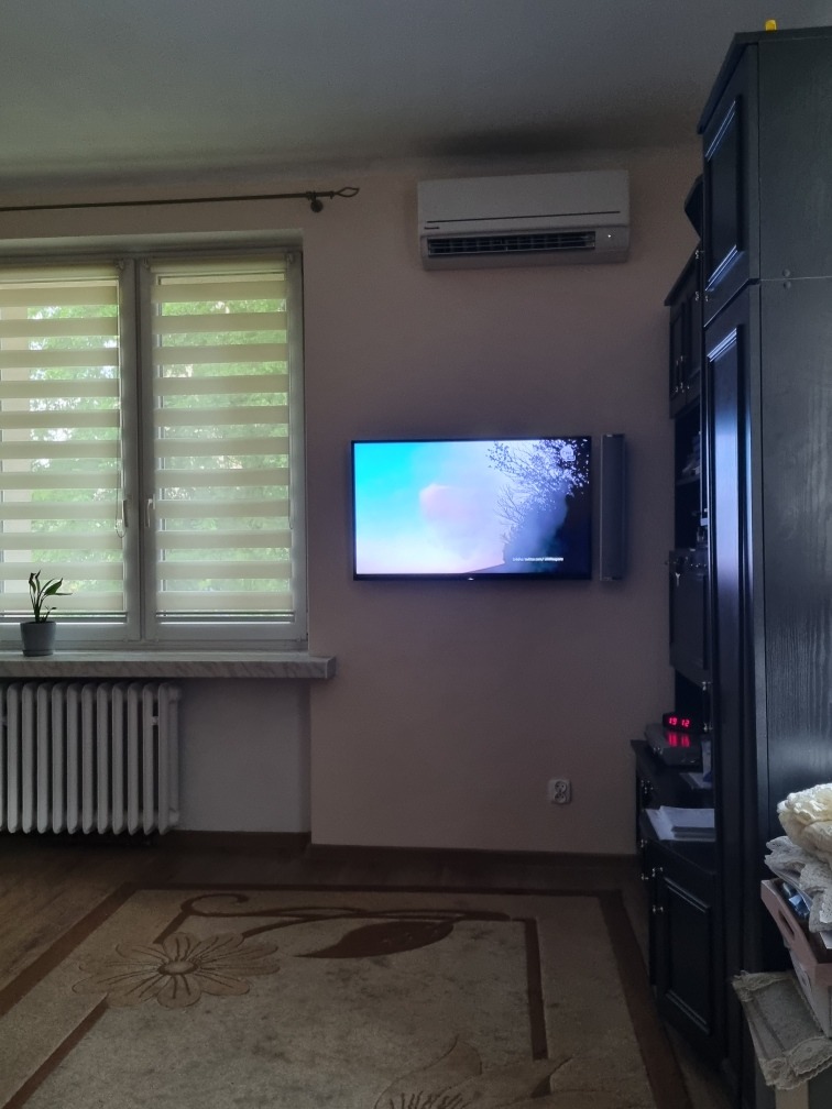 Klimatyzator ścienny zamontowany nad telewizorem w salonie z zasłoniętymi żaluzjami okiennymi i ciemnymi meblami.