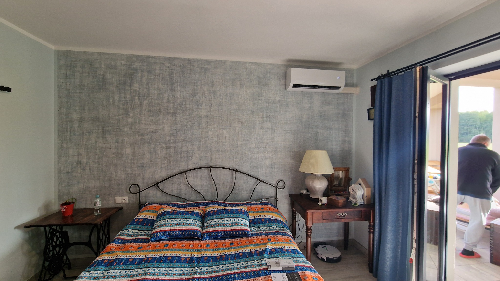 Jasny pokój z łóżkiem o metalowej ramie i kolorową narzutą. Na ścianie zamontowany klimatyzator. Obok łóżka stolik nocny z lampką i dekoracjami. W tle widoczne otwarte drzwi balkonowe i osoba...