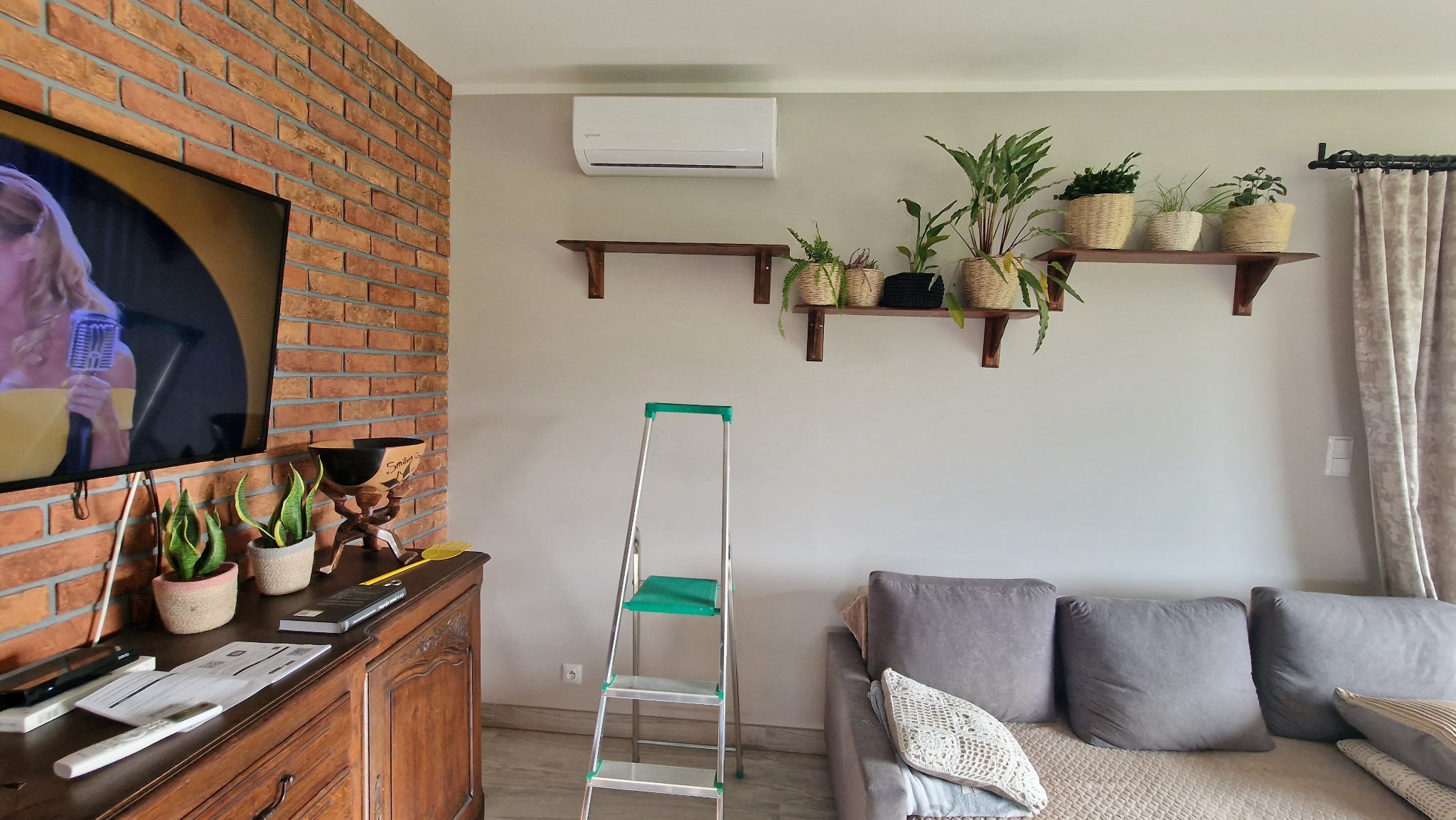 Wnętrze salonu z zamontowaną klimatyzacją ścienną; widoczna drabina malarska, drewniane półki z roślinami doniczkowymi, telewizor z ceglaną ścianą w tle oraz szara sofa.