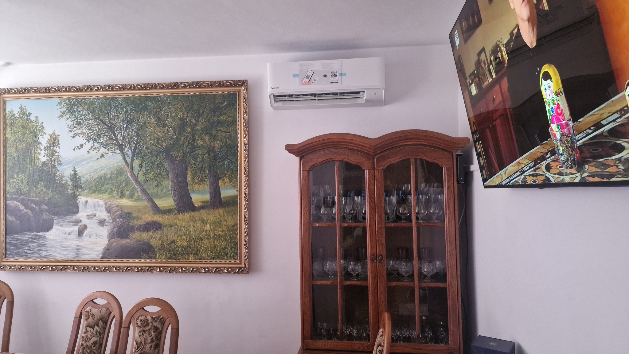 Biały klimatyzator Panasonic zamontowany na ścianie nad drewnianą witryną z kieliszkami, obok obrazu w złotej ramie przedstawiającego krajobraz z wodospadem i telewizora pokazującego odbicie wnętrza.