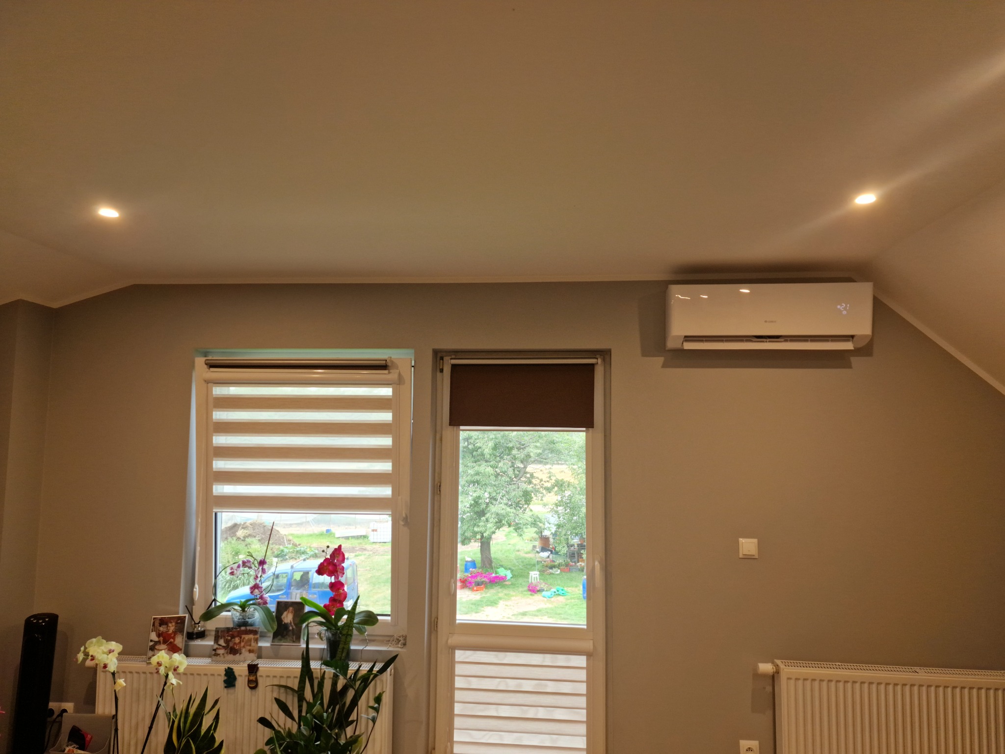 Biały klimatyzator ścienny zamontowany w pokoju z oknami, żaluzjami i grzejnikiem, widok na ogród.