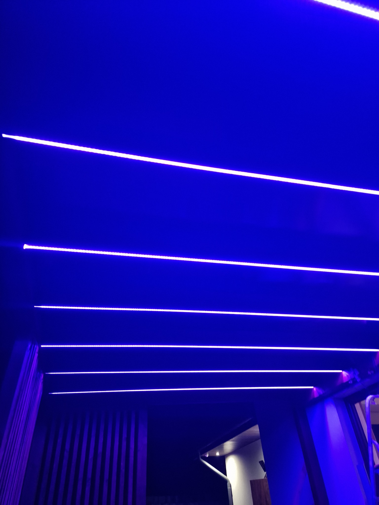 Nowoczesne oświetlenie LED w kolorze niebieskim, równoległe linie na suficie, widoczne elementy konstrukcyjne budynku.