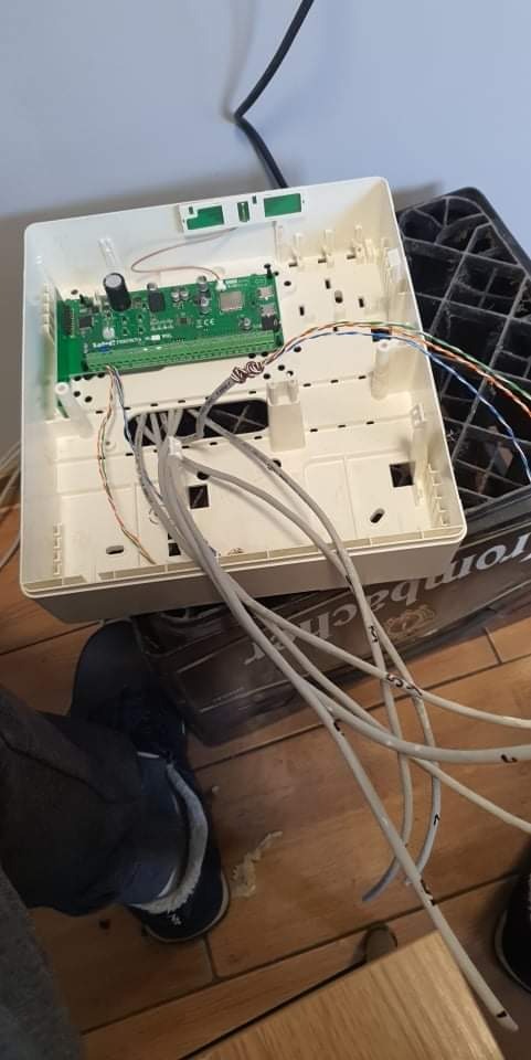 Otwarta biała skrzynka z zieloną płytką drukowaną i podłączonymi szarymi kablami na drewnianej podłodze, obok czarnej skrzynki z tworzywa sztucznego.