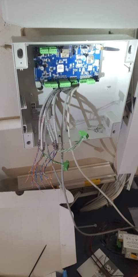 Otwarta skrzynka sterownicza z niebieską płytą główną, widoczne liczne kable podłączone do zielonych złączek, instalacja w trakcie montażu.