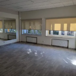 Puste, wyremontowane pomieszczenie z szarymi panelami podłogowymi, grzejnikami pod oknami z beżowymi roletami i lustrem na ścianie.