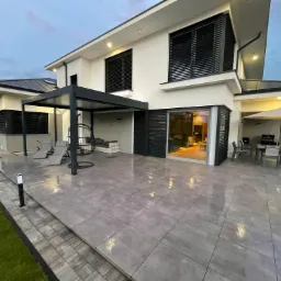 Nowoczesny dom jednorodzinny z płaskim dachem, dużymi oknami z żaluzjami zewnętrznymi i tarasem wyłożonym szarą płytką, widok z ogrodu po deszczu.