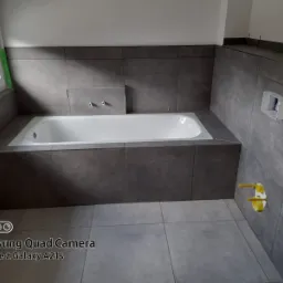 Biała wanna obudowana szarymi płytkami w łazience w Ząbkowicach Śląskich, widoczne instalacje wodne na ścianie i zabezpieczone okno.