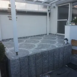 Wykończony taras z szarych płytek z dekoracyjnym wzorem w kształcie półokręgów, otoczony niskim murkiem z szarego kamienia, zadaszony konstrukcją z białych belek.