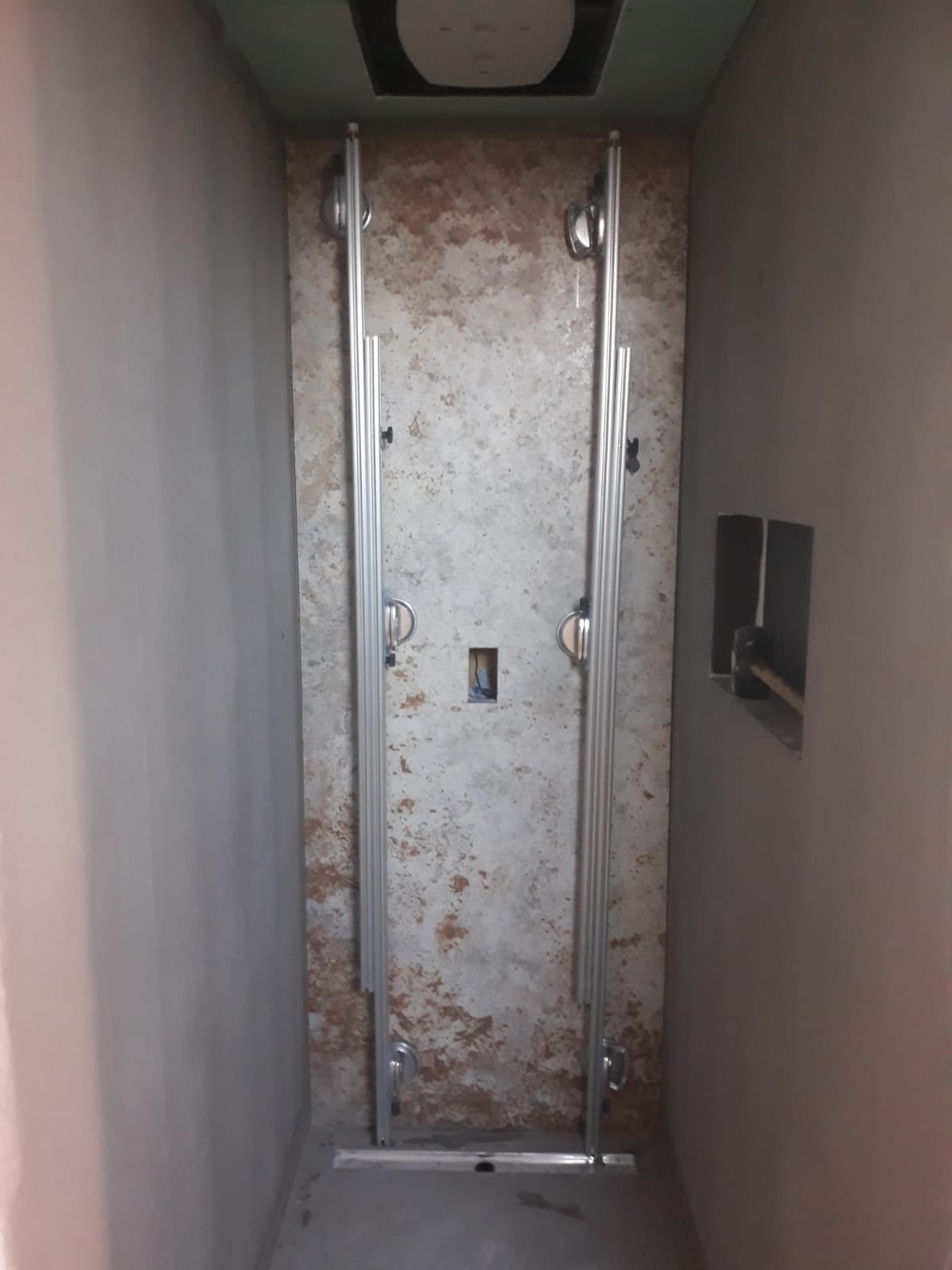 Przygotowanie do montażu kabiny prysznicowej: metalowe profile mocujące na ścianie z surową fakturą, widoczne instalacje w ścianie, surowy stan wykończenia pomieszczenia.