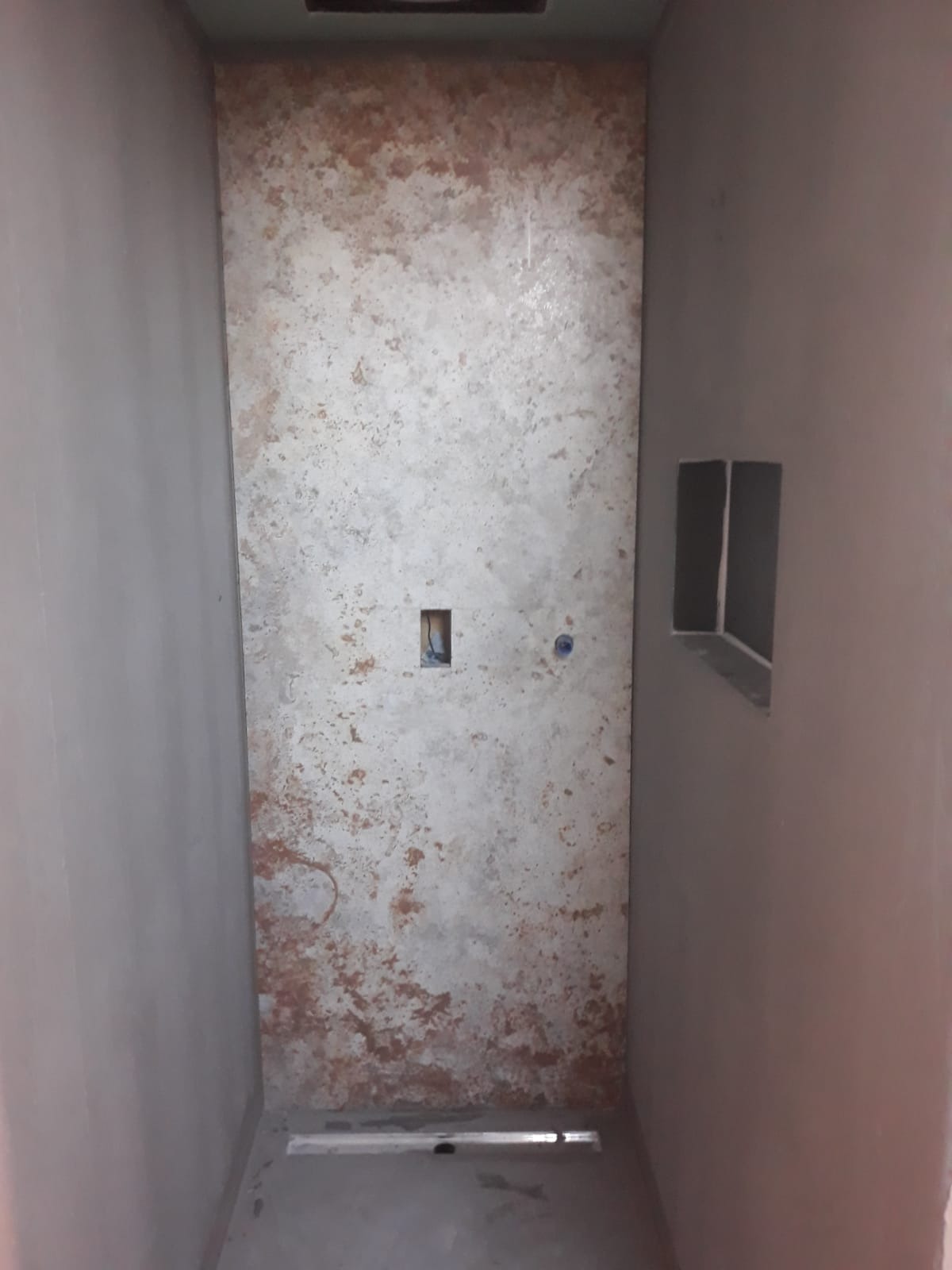 Surowa łazienka w trakcie remontu: ściana z efektem rdzy, otwór na gniazdko, wnęka w ścianie, odpływ liniowy w podłodze.