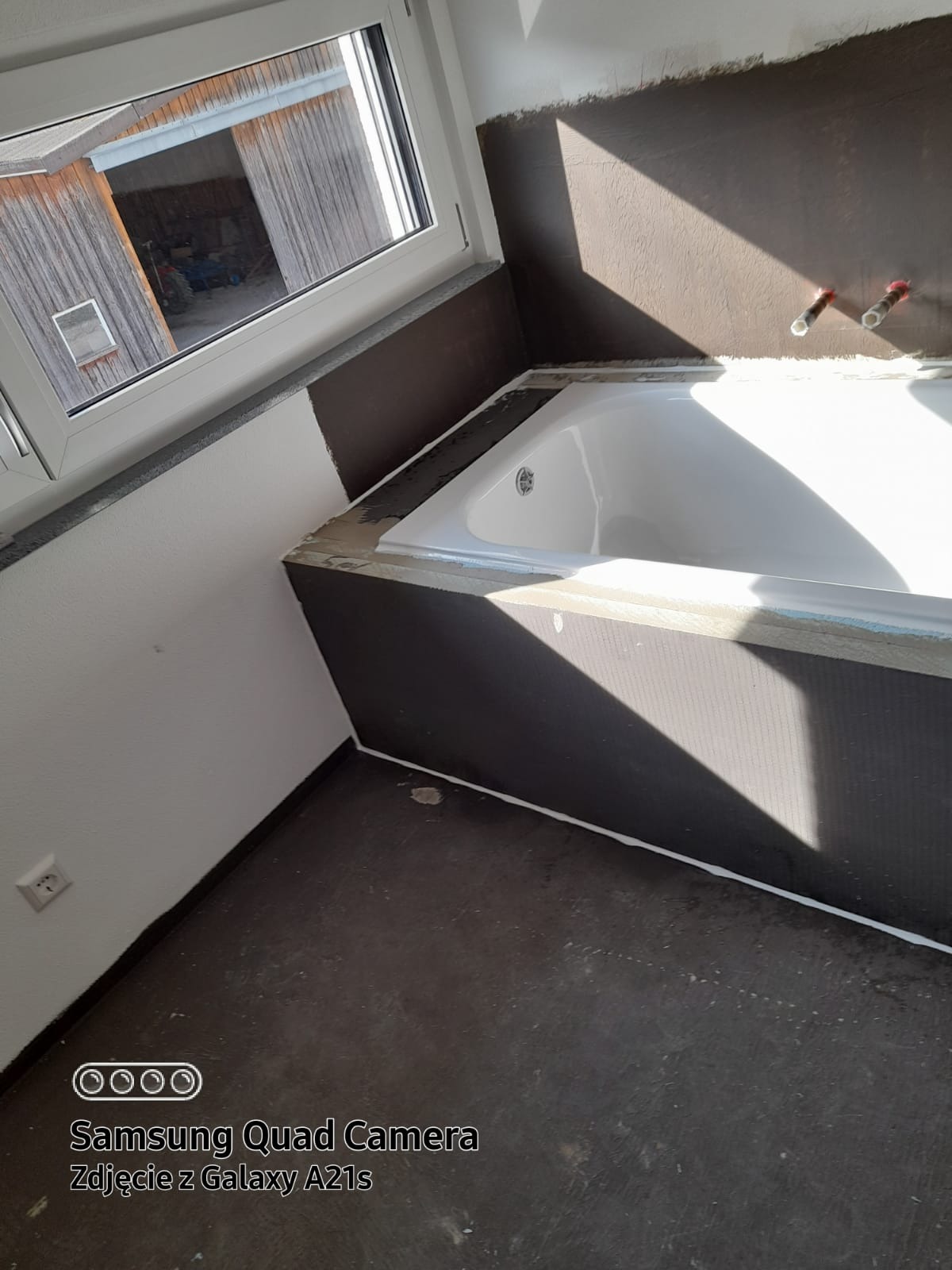 Biała wanna w trakcie montażu, widoczne przygotowane ściany z brązowym gruntem, okno z widokiem na budynek gospodarczy, surowy stan podłogi.