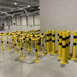 Żółte bariery ochronne z czarnymi pasami w hali magazynowej, widoczne filary konstrukcyjne i instalacje pod sufitem.